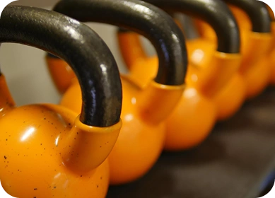Zbliżenie na kilka jasnopomarańczowych kettlebells z czarnymi uchwytami ułożonych w rzędzie na stojaku. Obraz ten oddaje poczucie siły i skupienia na sprawności fizycznej.