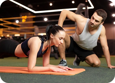 Kobieta znajduje się w pozycji deski na pomarańczowej macie w sali gimnastycznej, a mężczyzna w stroju sportowym kuca obok niej, udzielając jej wskazówek. Otoczenie jest dobrze oświetlone i wyostrzone.