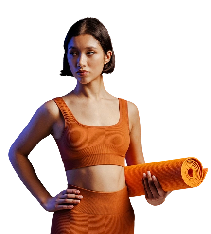 Kobieta w pomarańczowym stroju sportowym trzyma pod pachą zwiniętą matę do jogi na czarnym tle. Patrzy w bok z pewnym siebie wyrazem twarzy.